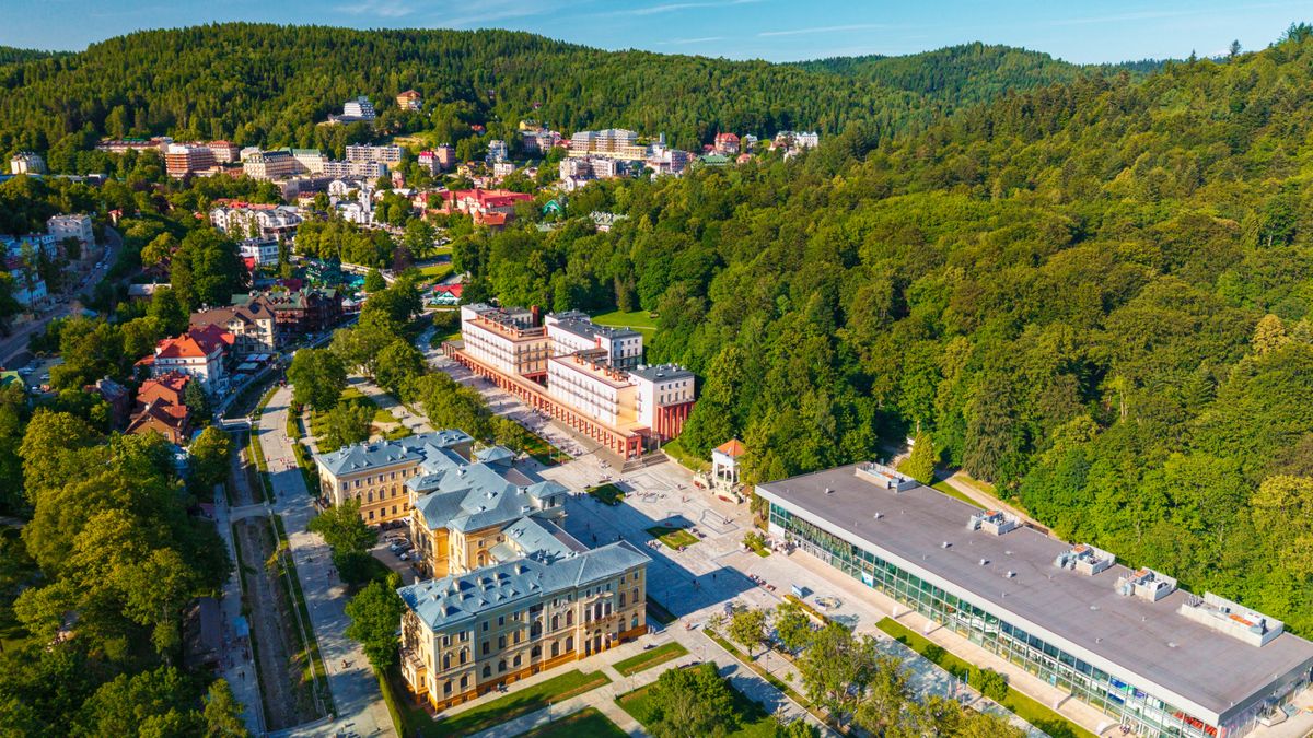 Widok z lotu ptaka na centrum Krynicy-Zdroju. Widoczny jest zielony deptak, zabytkowe i nowoczesne budynki uzdrowiskowe oraz Główna Pijalnia Wód, otoczone zalesionymi wzgórzami.