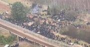 Setki  migrantów z plecakami i śpiworami przy granicy z Polską. Obok uzbrojeni ludzie w mundurach.