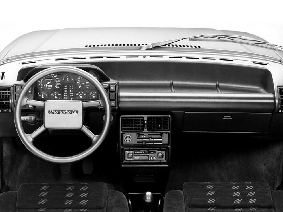Fiat Uno Turbo i.e.
Trzeba przyznać, że Włosi zawsze umieli w małe auta – zaczynając od modelu 500, przez Pandę, aż po Uno – Europejski Samochód Roku 1984, którego kolejna generacja produkowana była w latach 1995-2002 w tyskim zakładzie Fiata. Nas rzecz jasna najbardziej interesuje wersja Turbo, która przyspieszała do setki w zaledwie 8,3 sekundy. Mogła mieć cyfrowe zegary, podobnie jak zwykły model. Jednak tym razem chciałem się skupić na "zwykłym" kokpicie.
Jesteście fanami zwykłych zegarów? Właśnie trafiliście do raju, bo w Uno Turbo jest ich aż 7. Prędkościomierz, obrotomierz, wskaźnik paliwa, temperatura cieczy, temperatura oleju, ciśnienie oleju i na deser – wskaźnik doładowania. Dobrze, że wszystko się zmieściło. Aha, a gdyby ktoś zapomniał, że jedzie Uno Turbo z elektronicznym wtryskiem, to kierownica z uroczo cienkimi poprzeczkami, wyglądającymi jak nogi pająka, od razu o tym przypomni.