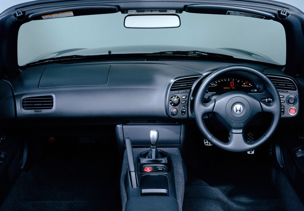 Honda S2000
Wydawać by się mogło, że Honda S2000 jest autem jeszcze stosunkowo młodym, ale co jeśli powiem, że jej debiut odbył się… 22 lata temu? Szmat czasu, a kręcący się do blisko 9000 obr/min, 240-konny wolnossący silnik do dziś wywołuje dreszcze, gdy mignie na ulicy. Japoński roadster zestarzał się świetnie nie tylko z zewnątrz, ale także w środku.
Minimalizm panuje tu wszędzie, poza miejscem kierowcy – wszystkie funkcje rozmieszczono blisko kierownicy, tak, by możliwie najbardziej ograniczyć ruchy ręką – pokrętła nawiewów z prawej, obsługa radia z lewej, podobnie jak duży czerwony przycisk do odpalania. A skoro o radiu mowa, to jest schowane za osłoną przed dźwignią zmiany biegów. A całość dopełniają elektroniczne zegary w stylu F1. Nie trzeba być fanem JDM-u, żeby się w tym zakochać.