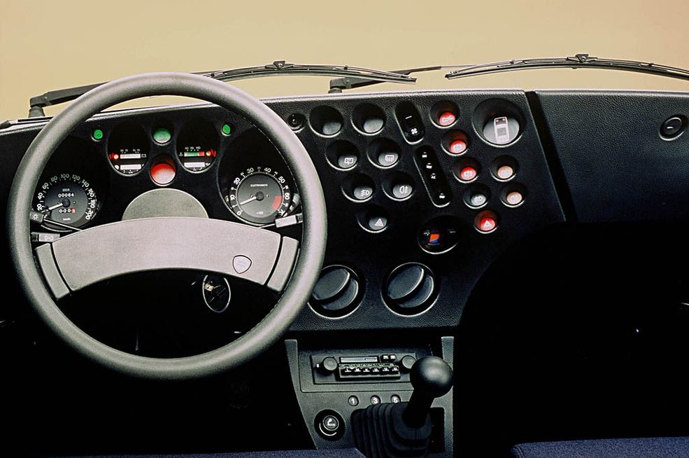 Lancia Beta Trevi
Choć 4-drzwiowy sedan Trevi nie ocieka włoskim stylem, a jego pudełkowate nadwozie można wręcz określić mianem nudnego, tak wnętrze jest absolutnie niepowtarzalne. I może trudno w to uwierzyć, ale jedno i drugie projektowała ta sama osoba. Ze względu na liczne otwory, zarówno na konsoli środkowej, jak i za kierownicą, w których umieszczono zegary, lampki kontrole, check panel oraz wszelkie możliwe przełączniki i przyciski, deskę rozdzielczą porównywano do szwajcarskiego sera. Były one jednak osadzone na tyle głęboko i zwrócone w stronę kierowcy, że pasażer ledwo był w stanie obsłużyć konsolę nawiewu. Oprócz tego Lancia Trevi w wersji VX była pierwszym powojennym, seryjnie produkowanym samochodem wyposażonym w mechaniczną sprężarkę (Rootsa).