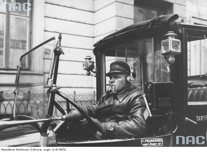 1935
Warszawski taksówkarz palący fajkę za kierownicą auta wyposażonego w zwijany brezentowy dach nad miejscem kierowcy i oddzielną kabiną pasażerską. Pojazd wyposażony był radioodbiornik ze słuchawkami. Na zdjęciu widoczny także klakson oraz boczne lampy elektryczne.
