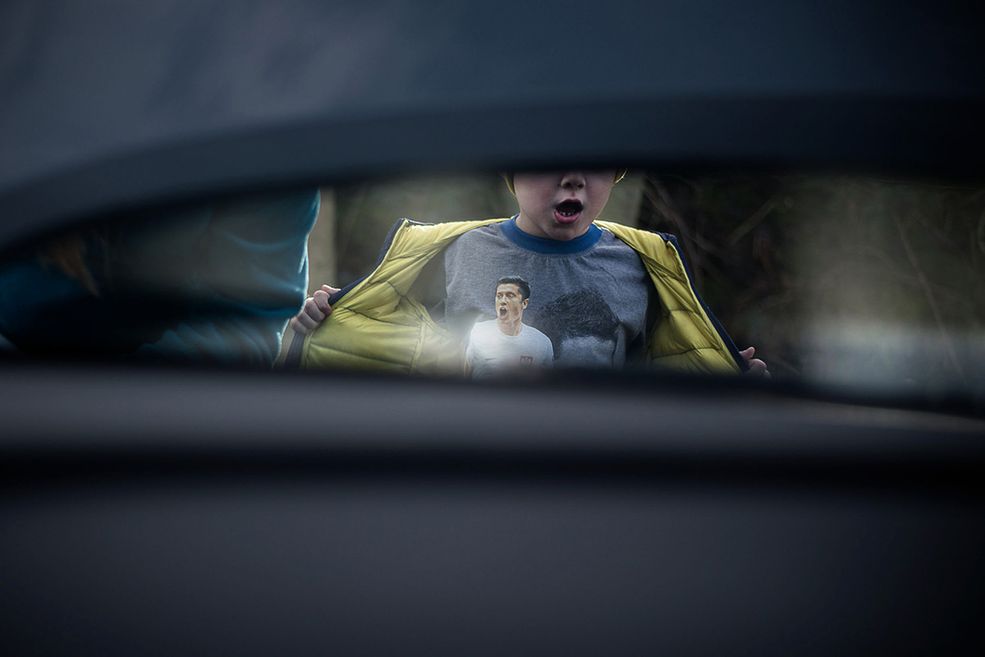 Zdjęcie sprawia wrażenie podpatrzonego momentu przez ciasną szczelinę ulicznej zabudowy. Wszyscy, którzy otarli się o piłkę nożną, mają zapewne w głowach obrazy ukazujące radość piłkarzy. To powtarzające się uniwersalne gesty i jeden z nich, zapewne powielony, jest również na koszulce chłopca. Jest to gest niemal identyczny, jednak wykonywany w zupełnie innym znaczeniu niż ten piłkarski – jakby było chłopcu za gorąco albo po prostu chciał pobawić się kurtką. Otwarta buzia potęguje to wrażenie, ponieważ znowu nawiązuje do koszulki. Ta pozornie zwykła uliczna scena przywołała mi wspomnienia sukcesów reprezentacji podczas niedawnego Euro oraz tysiące młodych dzieciaków, którzy chcą być jak Lewandowski.