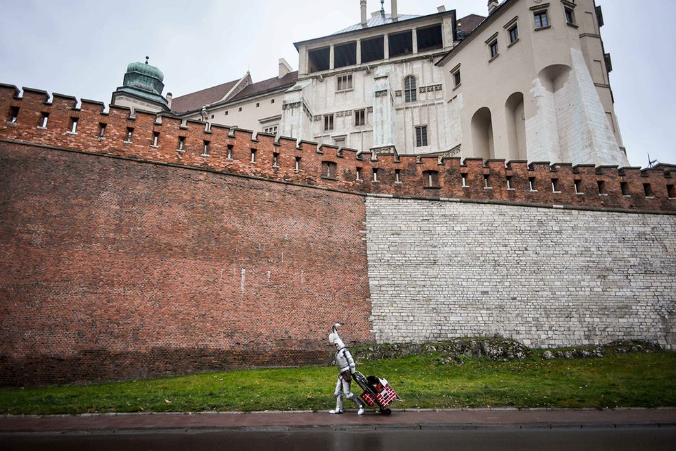 Rycerz pod Wawelem łączy czasy minione (zbroja) i teraźniejsze (wózek z gadżetami). Ten dualizm widoczny jest też na ścianie, gdzie kolory nawiązują do biało-czerwonej polskiej flagi. Faktura cegieł powielona jest na wózku. Szeroki prosty kadr podkreśla monumentalność Wawelu. Rycerz wraca z pracy – wyobrażam go sobie jadącego w tej zbroi nowoczesnym tramwajem, wchodzącym na nowoczesne zamknięte osiedle i znikającego w domowym zaciszu, odkładając hełm na wieszak, a miecz w pochwie do stojaka na parasole. Jutro czeka go kolejny dzień pracy.