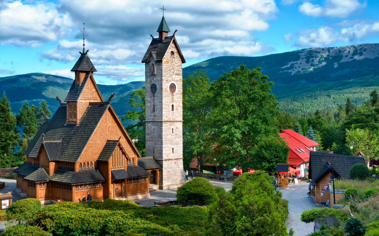 Zabytkowy, drewniany kościółek Wang (Ewangelicki Kościół Górski Naszego Zbawicielaw) Karpaczu o unikalnej architekturze nordyckiej, z wysoką, kamienną dzwonnicą stojącą tuż obok. Całość otacza zadbany ogród i zielone drzewa, a w tle rozciąga się majestatyczna panorama zalesionych szczytów Karkonoszy pod błękitnym niebem z białymi chmurami.