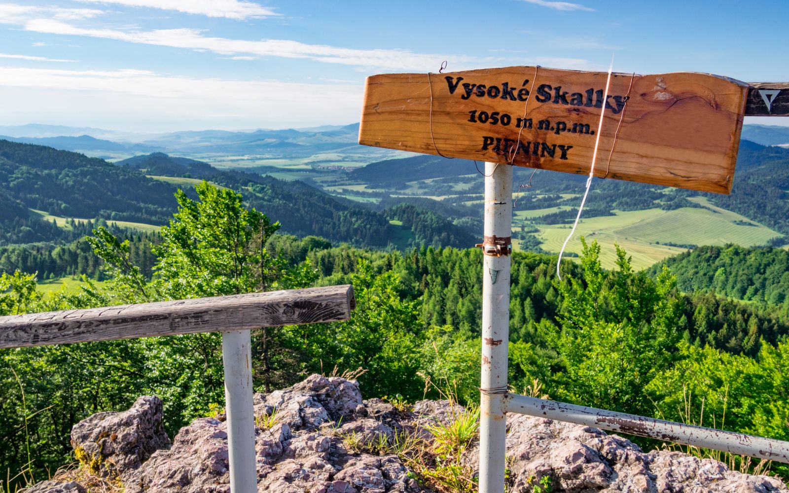 Widok z pienińskiego szczytu. Na pierwszym planie po prawej stronie stoi metalowy słupek z drewnianą tablicą z napisem „Vysoké Skalky 1050 m n.p.m. PIENINY”. W tle rozciąga się rozległa panorama  wzgórz, lasów i pól pod błękitnym niebiem z delikatnymi chmurami.