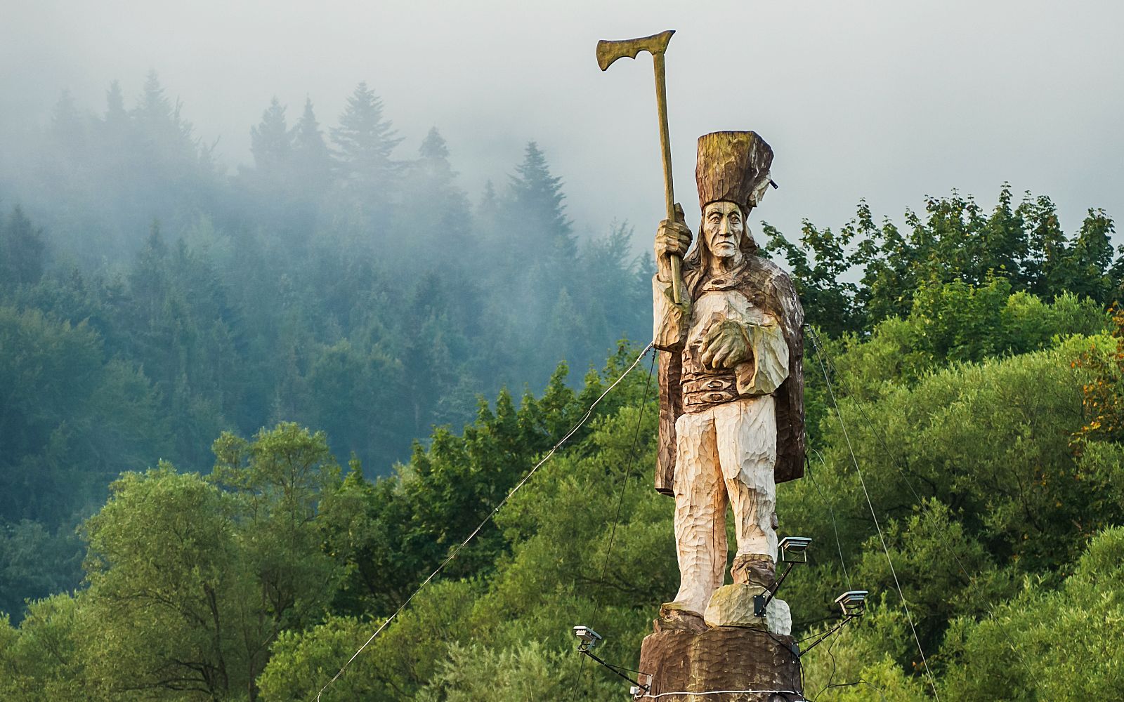 Zbliżenie na monumentalną, rzeźbioną w drewnie figurę górala w tradycyjnym stroju i wysokiej czapie, trzymającego w uniesionej dłoni dużą ciupagę. Rzeźba Górala na Kotuńce stoi na podwyższeniu, otoczona reflektorami i linami montażowymi. W tle rozciąga się gęsty, zielony las częściowo spowity poranną mgłą.