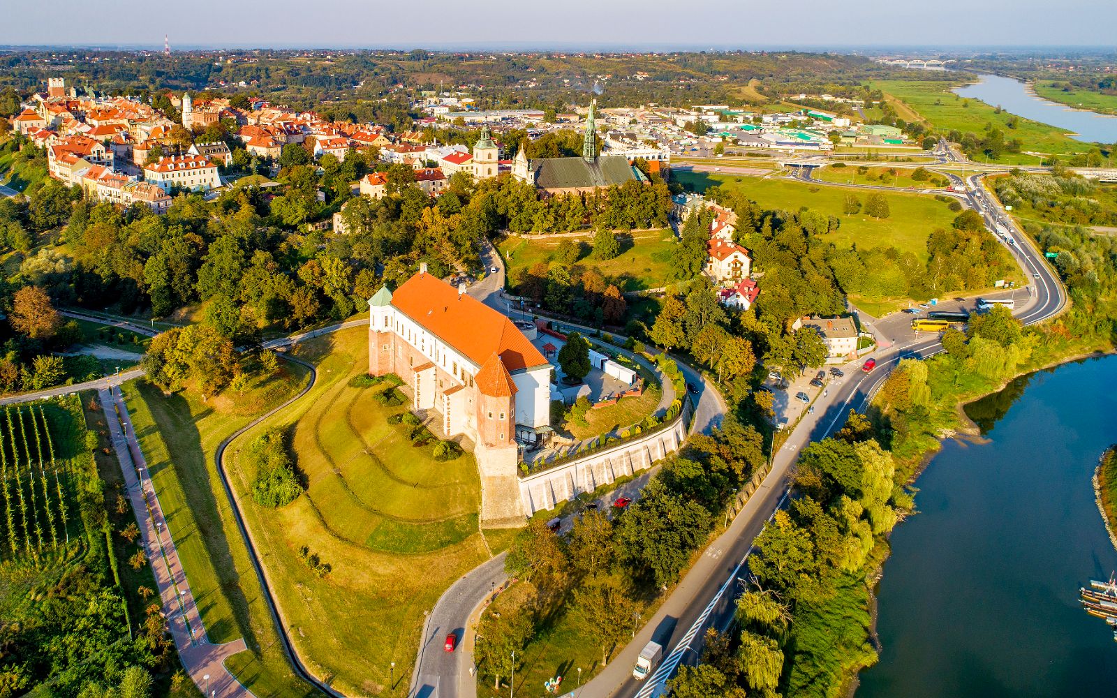 Widok z lotu ptaka na Sandomierz w słoneczny dzień. W centrum kadru na zielonym wzgórzu wznosi się renesansowo-gotycki Zamek Królewski o białych ścianach i dużym dachu z czerwonej dachówki. Po prawej stronie wije się koryto rzeki Wisły, a z lewej strony w tle widać gęsto zabudowaną, zabytkową starówkę z licznymi kamienicami o czerwonych dachach i wieżami kościołów pośród zieleni drzew.