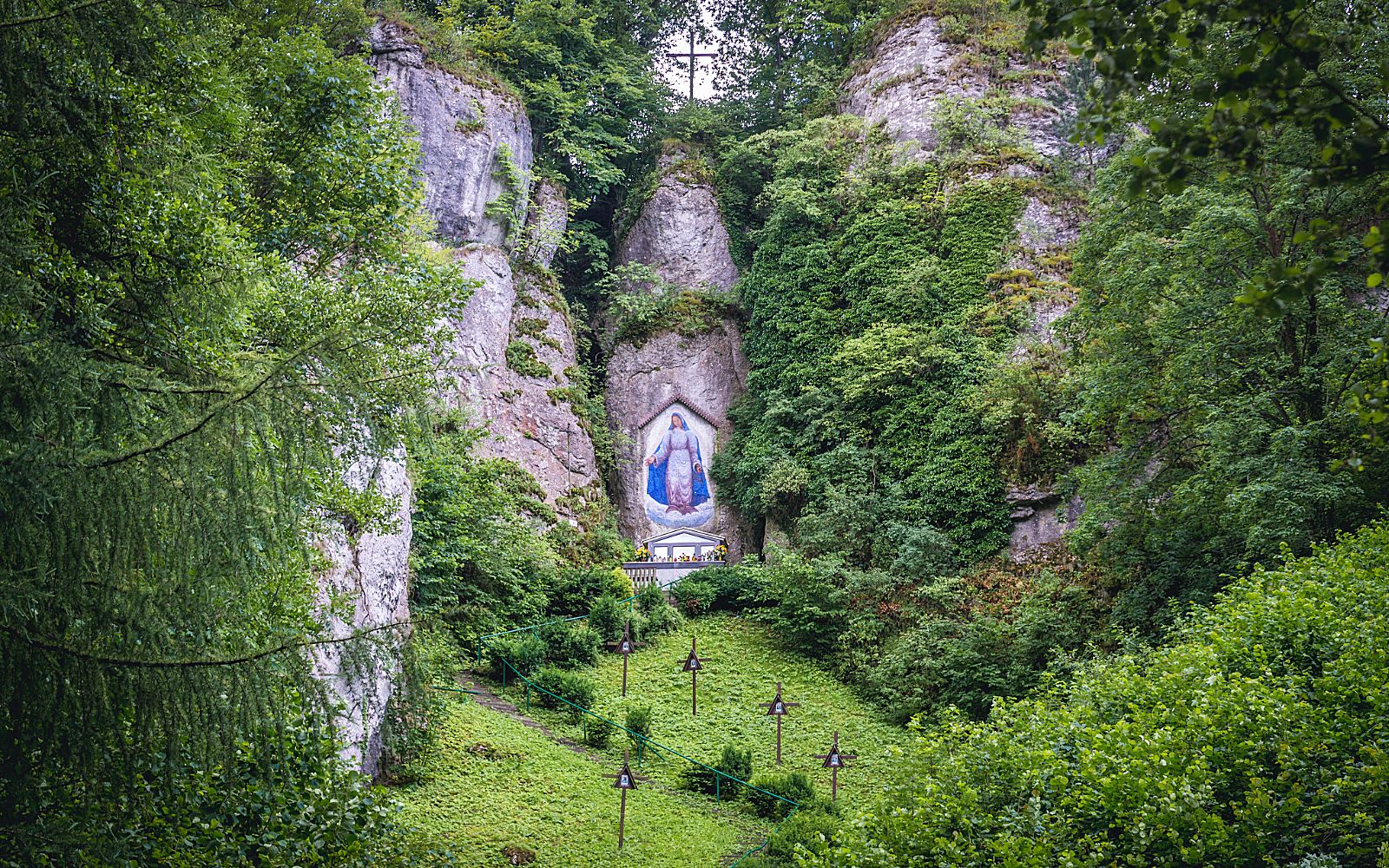 Naskalny obraz Matki Bożej w niebiesko-białej szacie, namalowany we wnęce wysokiej, wapiennej skały pokrytej bluszczem. Poniżej obrazu znajduje się mały, polowy ołtarz, do którego prowadzi ścieżka po zielonym zboczu wzgórza. Wzdłuż ścieżki ustawione są drewniane krzyże stacji Drogi Krzyżowej, a całe ujęcie otaczają gęste, zielone korony drzew.