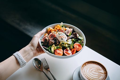 Close up of woman's hand holding a bowl of fresh beef cobb salad, serving on the dining table. Ready to enjoy her healthy and nutritious lunch with coffee. Maintaining a healthy and well-balanced diet. Healthy eating lifestyled3sign